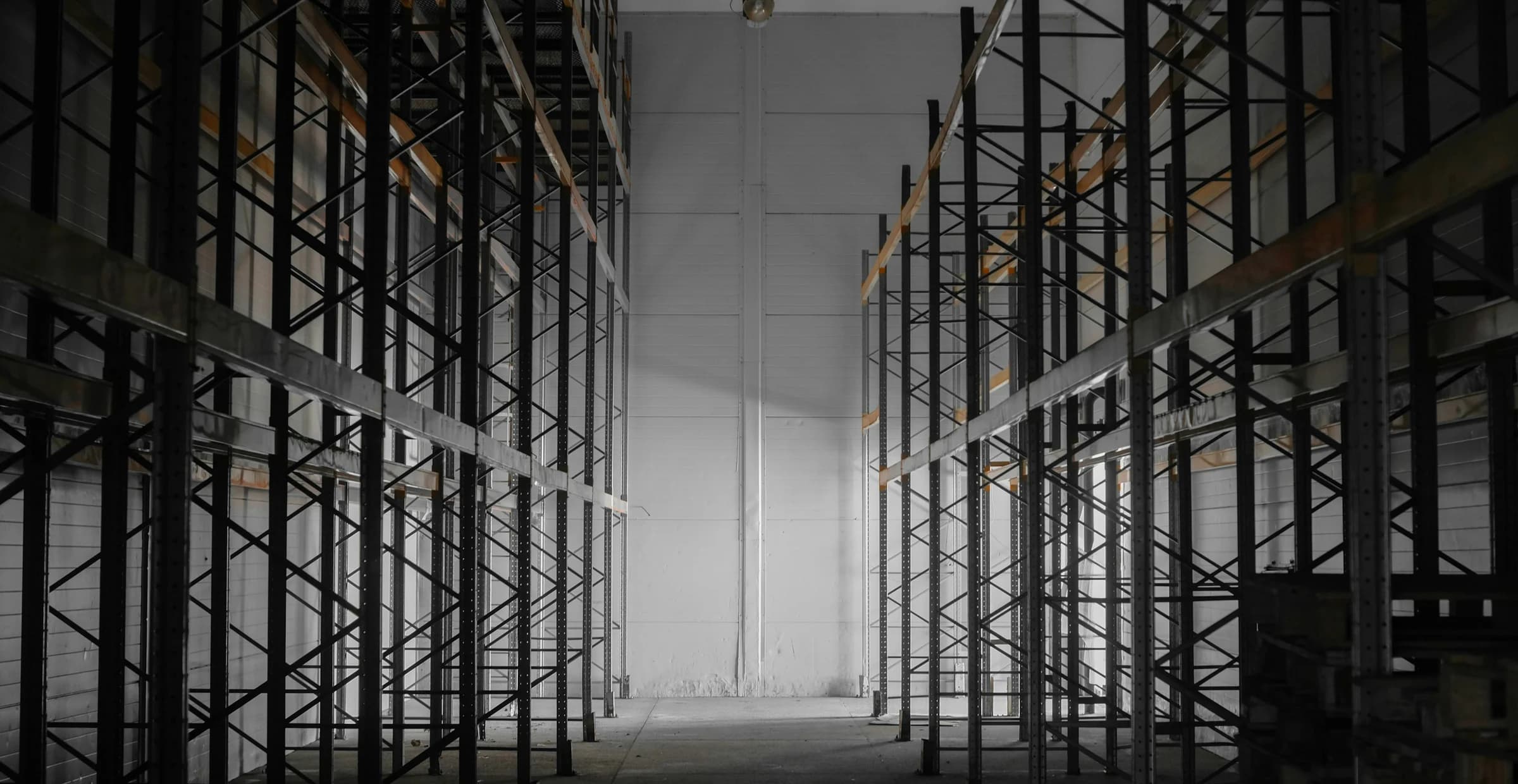 A tall aisle of empty pallet racking in a warehouse, receding toward a lit far wall